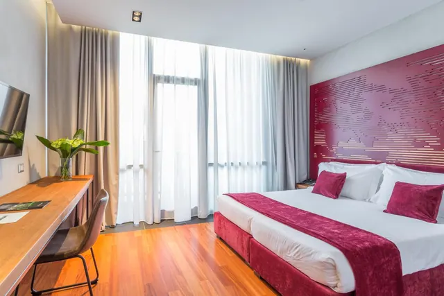 A bedroom with a double bed under a magenta headboard, a wooden desk with a chair, a wall-mounted television and floor-to-ceiling windows behind sheer curtains.