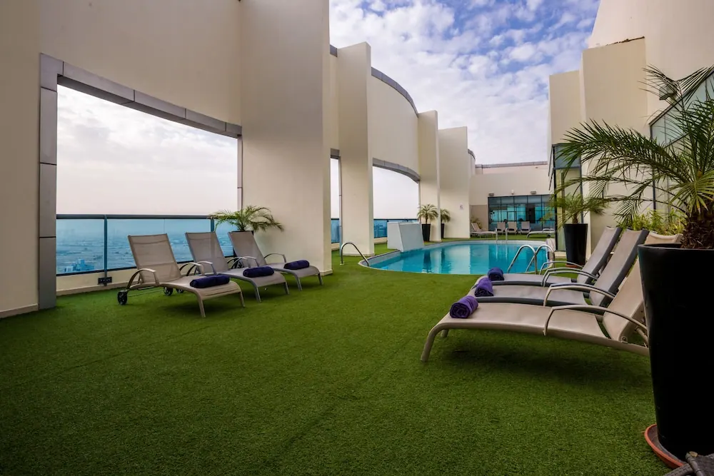 A small rooftop pool surrounded by artificial grass and sun loungers with purple towels, with a glass railing and hazy distant buildings visible on the left.