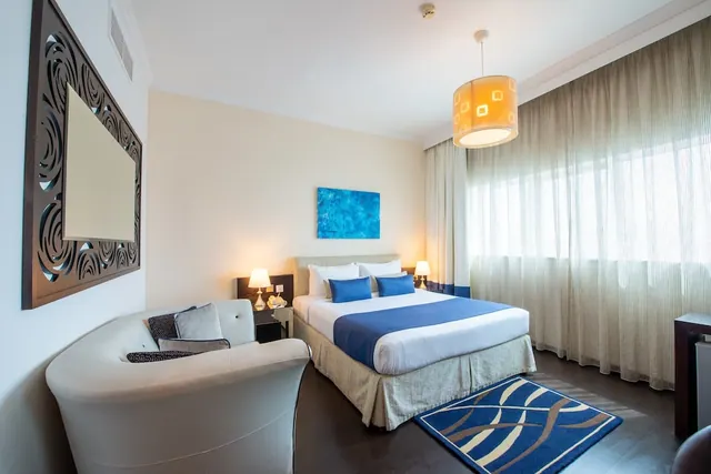 A hotel bedroom with a made double bed with blue accents, a white armchair, bedside lamps, a pendant light and a window behind sheer curtains.