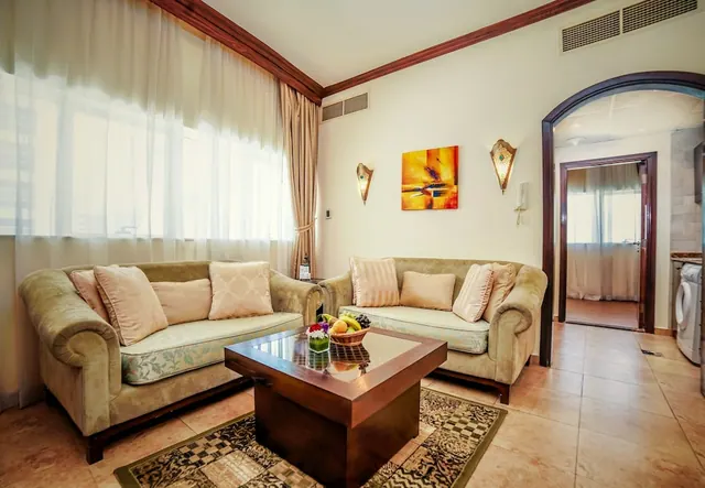 A living room with two beige sofas facing each other across a glass-topped wooden coffee table holding a fruit bowl, with sheer curtains at a window and an arched doorway to another room.