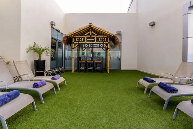 An outdoor terrace with artificial grass has sun loungers with purple towels and a bamboo gazebo with bar stools in front of a white wall.