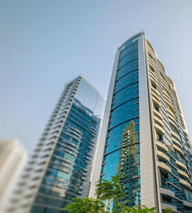 Low-angle view of two tall glass towers with curved balconies under a clear blue sky, with green palm foliage in the foreground.