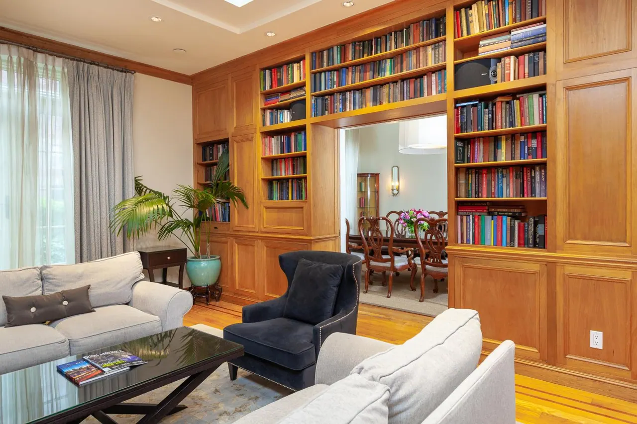 A living room with built-in wood bookshelves, two grey sofas, a navy armchair and a glass coffee table, with a dining room visible through the doorway.