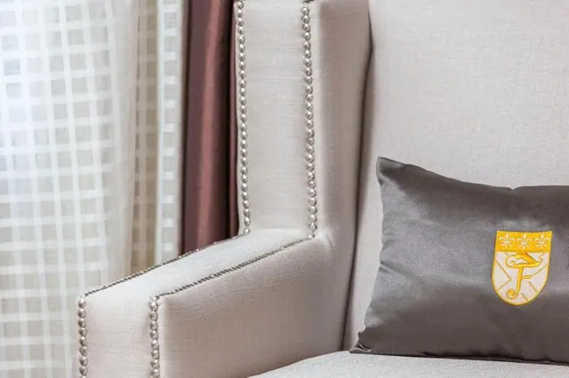 A close-up of a cream nailhead-trimmed armchair beside a grey satin cushion with a gold embroidered crest, with sheer checked curtains and a mauve curtain behind.