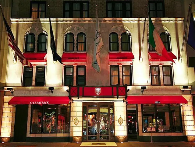 The illuminated night facade of a multi-storey building with red awnings, flags on the upper floors, and the name FITZPATRICK on the entrance canopy.