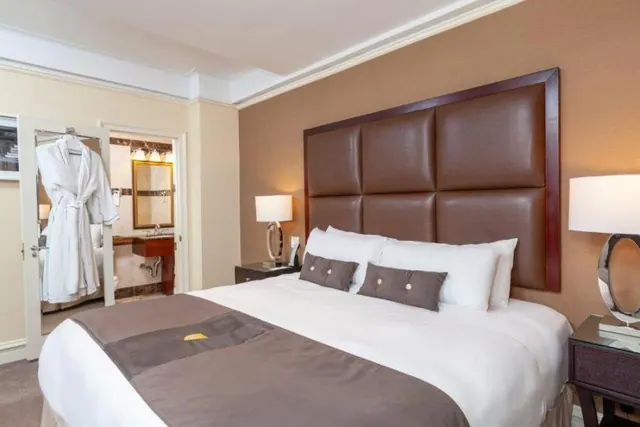 A king bed with a tufted brown leather headboard and white bedding sits beside lamps, with a white bathrobe hanging by an open door that leads to a bathroom.