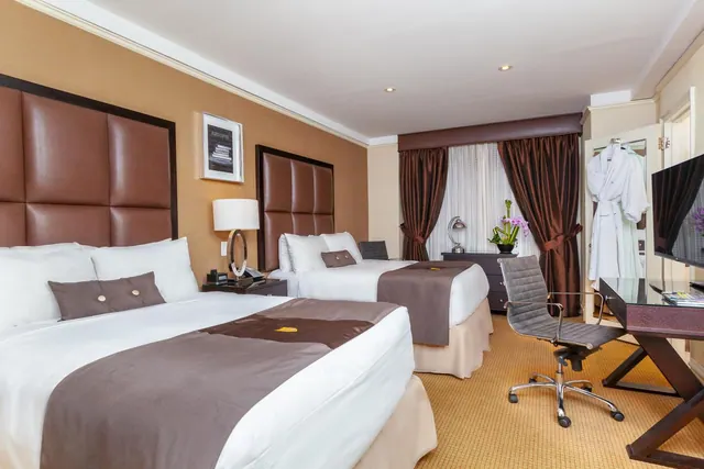 A hotel room with two double beds with brown leather headboards, a desk and chair, a flat-screen television, and a bathrobe hanging on the wardrobe door.