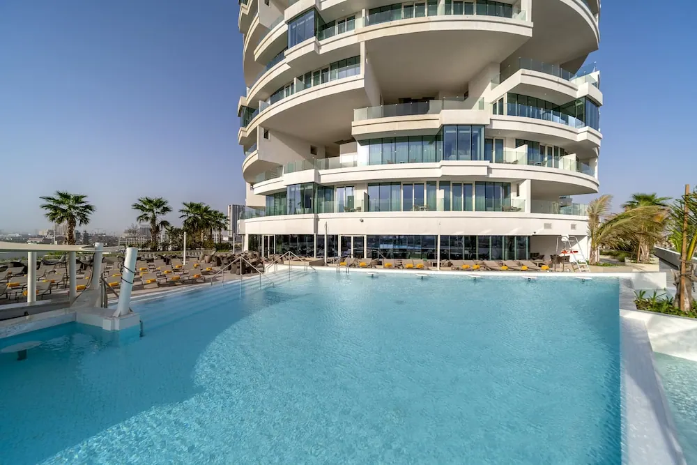 A large outdoor pool sits in front of a curved white residential tower with glass balconies, with rows of yellow sun loungers and palm trees beside it.