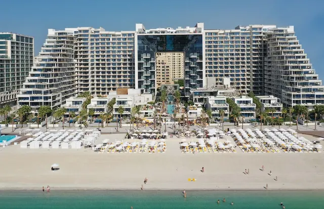 Aerial view of a high-rise hotel complex with a sandy beach, rows of white sun loungers and umbrellas, and turquoise sea in the foreground.