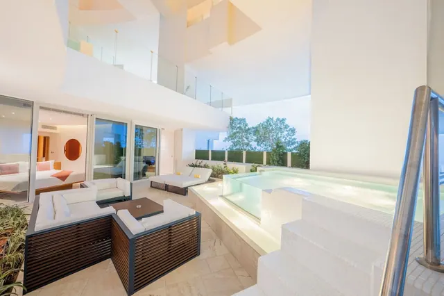 A terrace with a sunken plunge pool, white outdoor lounge seating and sun loungers, with a bedroom visible through glass doors.