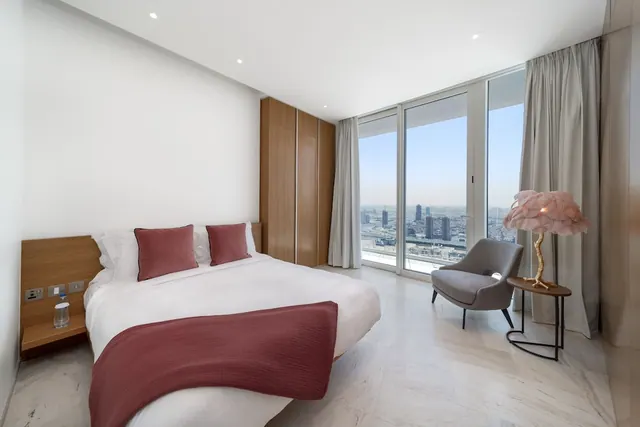 A bedroom with a made double bed, a grey armchair and floor-to-ceiling windows looking out over a city skyline.