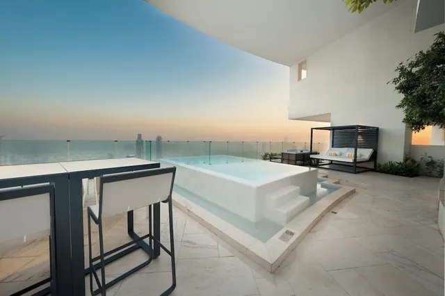 A private plunge pool and lounge seating on a terrace overlooking the sea and distant towers at dusk.