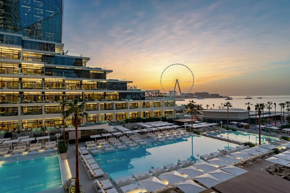 Sunset view of an outdoor pool with white-canopied sun loungers, a hotel building, and the Ain Dubai ferris wheel above the sea.