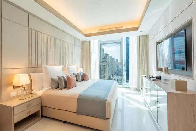 A bedroom with a made double bed, a wall-mounted television, and a large window overlooking high-rise towers.