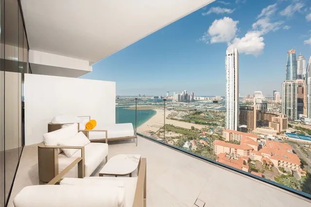 A furnished balcony with white lounge chairs and a glass railing overlooks a sandy beach, the sea and high-rise buildings.
