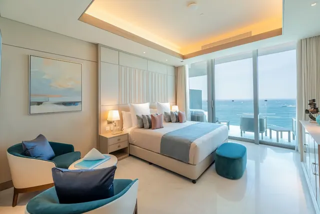 A bedroom with a king bed, two teal armchairs and a teal ottoman, with floor-to-ceiling windows looking out over the sea to a balcony.