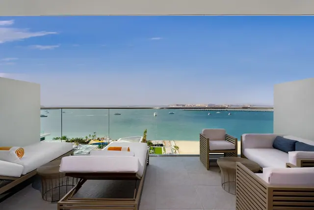 A furnished terrace with sun loungers and sofas overlooks the sea, a sandy beach, a pool and distant buildings.