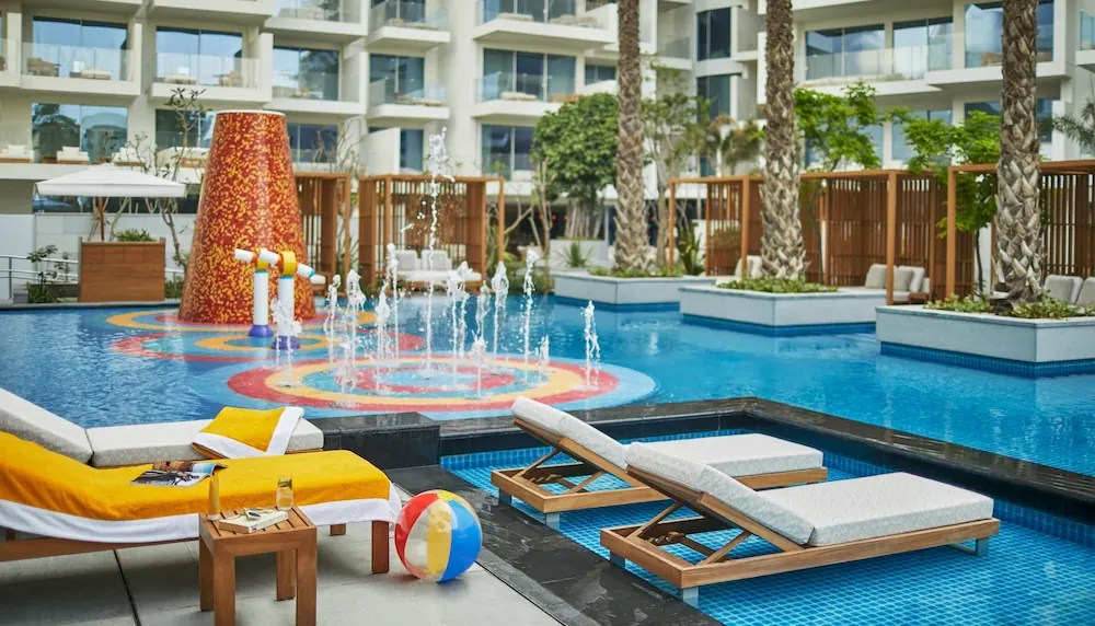 A large pool with a red and yellow water feature and jets, sun loungers with a yellow towel in the foreground, and palm trees beside a multi-storey building with balconies.