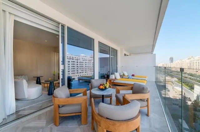A furnished balcony with teak armchairs, a round table holding a fruit bowl, yellow-cushioned daybeds and a glass balustrade overlooks a city street and residential buildings, with a white-furnished living room visible through the sliding doors.