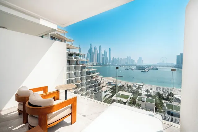 A white balcony with two wooden armchairs and a small round table overlooks a sea inlet, sandy beach, Dubai Marina towers and the Ain Dubai wheel.