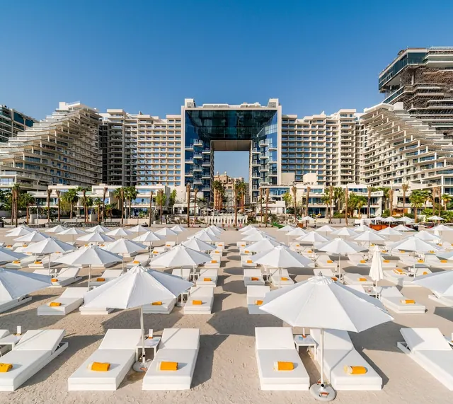 White umbrellas and loungers with orange towels stand on a sandy beach in front of large hotel towers under a clear blue sky.
