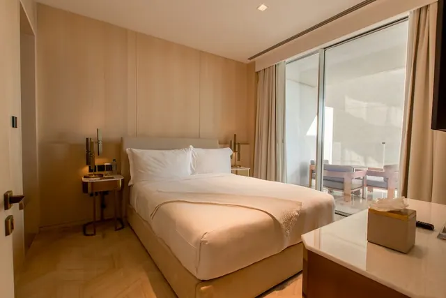 A bedroom with a double bed, bedside tables with lamps, and a sliding glass door opening onto a balcony with chairs.