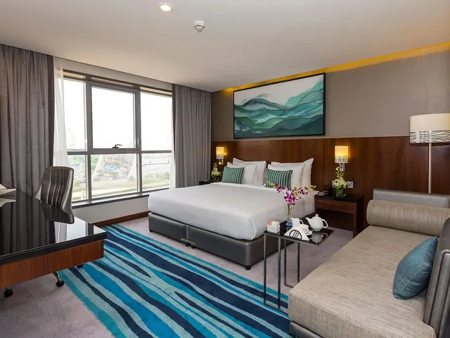 A hotel bedroom with a king bed, a grey sofa, a work desk, a tea tray on a side table, and a window looking out over buildings.