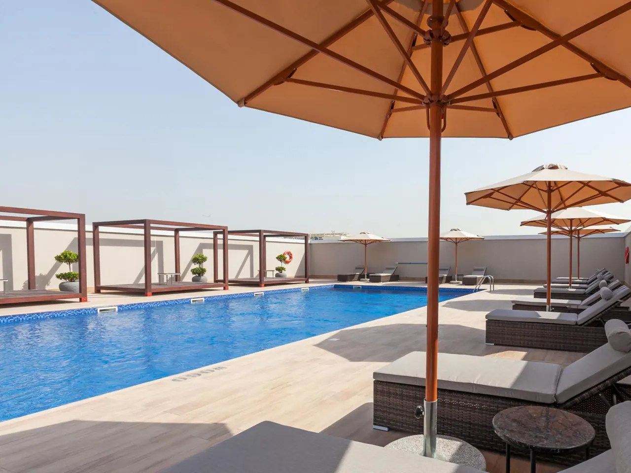 A rooftop swimming pool with sun loungers and parasols on a tiled deck, with wooden cabana frames and potted topiary along the far side.