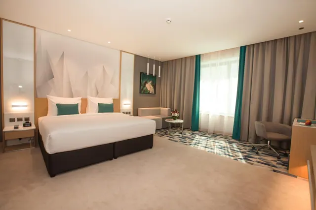 A hotel bedroom with a large bed dressed in white linen and teal cushions, a grey sofa and round side table by a curtained window, and a desk with a chair on the right.