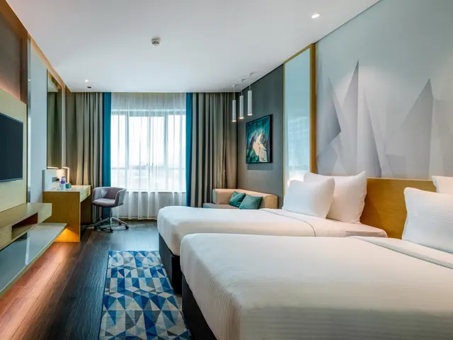 A hotel room with two beds, a sofa with teal cushions, a desk and chair, and a wall-mounted television.