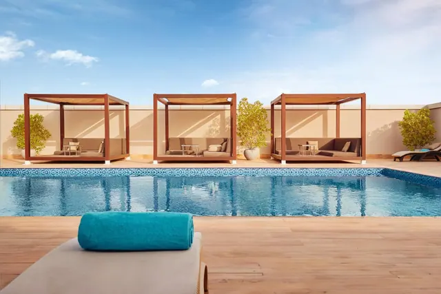 A tiled-edge swimming pool with three wooden canopied daybeds along the far wall and a rolled teal towel on a sun lounger in the foreground.