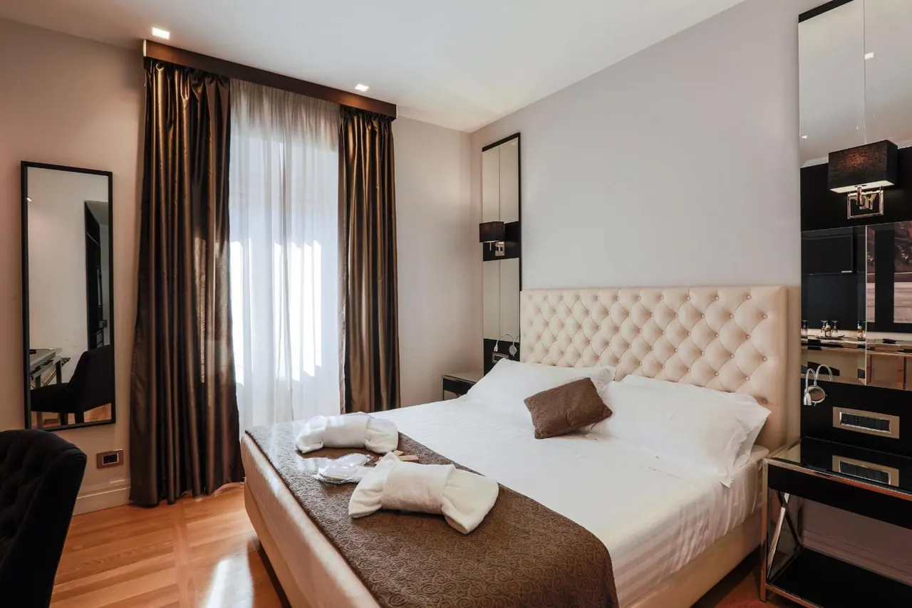 A bedroom with a double bed under a tufted cream headboard, a brown runner with two towel animals on the bed, dark curtains beside a sheer-curtained window, and a large mirror on the wall.
