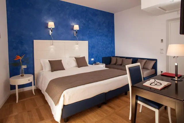 A hotel bedroom with a double bed in front of a blue textured wall, a sofa on the right, and a desk with an open book and glasses in the foreground.
