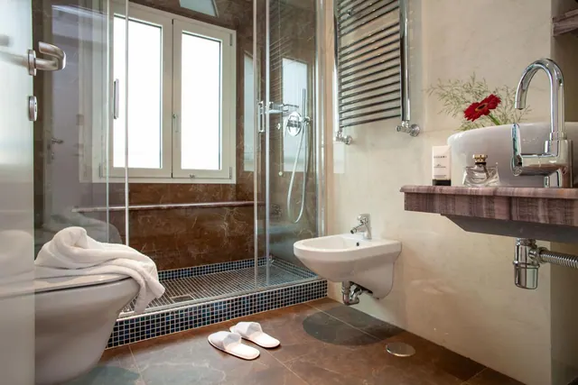 A bathroom with a walk-in glass shower, a wall-mounted bidet, and a vessel sink on a wooden shelf, with a folded towel on the toilet and white slippers on the floor.
