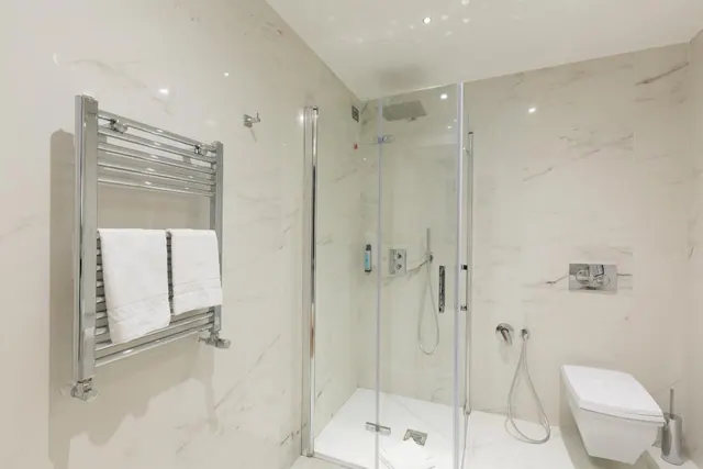 A white marble-tiled bathroom with a glass-walled walk-in shower, a heated towel rail holding white towels, and a wall-mounted toilet.