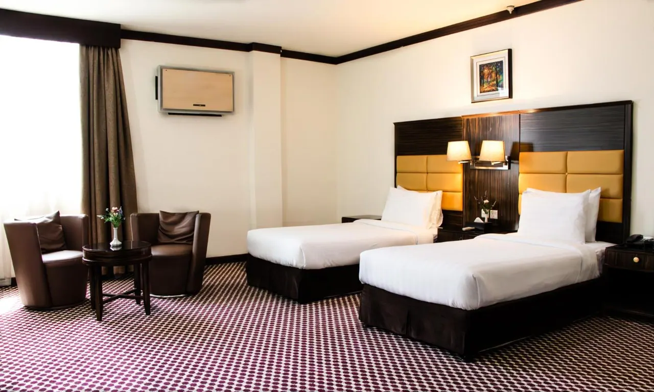 A hotel room with two single beds with tan padded headboards, two armchairs beside a small round table with flowers, and a wall-mounted television.