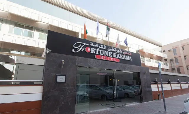 Street-level view of the Fortune Karama Hotel entrance, with a dark signboard, glass entrance doors, flags on the upper facade, and a parking sign on the pavement.