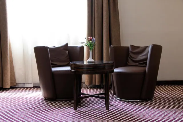 Two brown leather armchairs and a dark wood round side table with a vase of flowers stand in front of sheer and brown curtains on a patterned carpet.