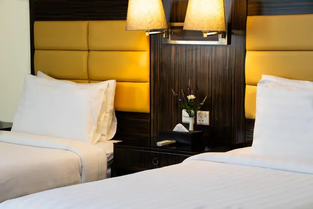Two single beds with white linen and yellow padded headboards sit either side of a dark wood nightstand holding a flower vase, tissue box and remote.