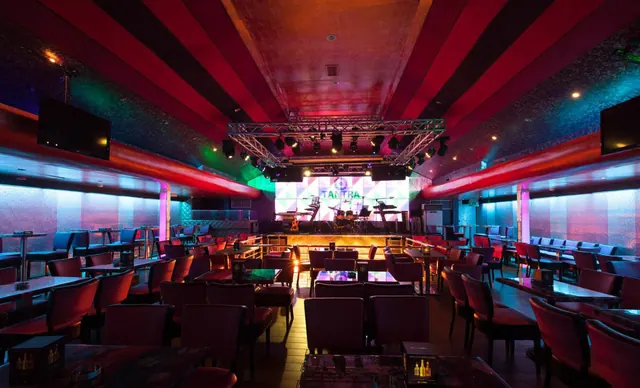Interior of a lounge with red and blue lighting, a stage with a screen showing the word TANTRA, and rows of chairs and tables.