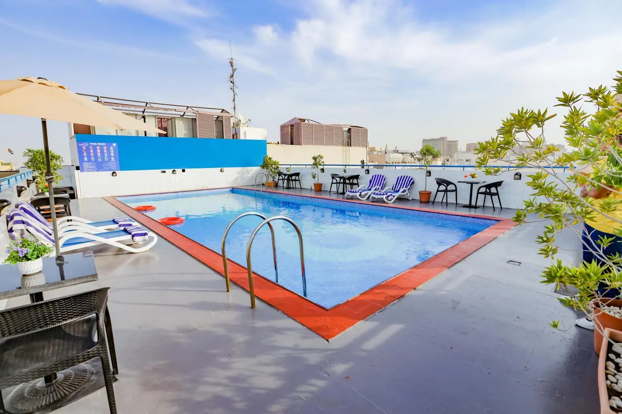A rooftop swimming pool with a red tiled edge, striped sun loungers, a cream umbrella and distant city buildings under a partly cloudy sky.