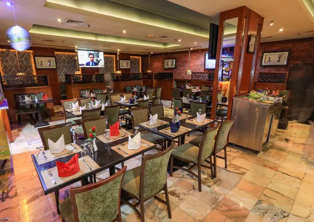 A dining room with rows of tables set with folded napkins and glassware, green upholstered chairs, a marble floor and a wall-mounted television.