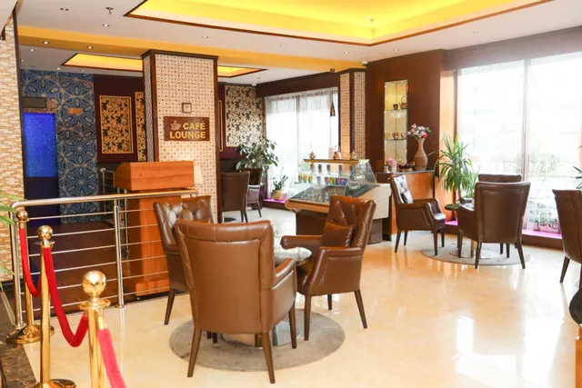 A lounge seating area with brown leather armchairs and round tables, a Café Lounge sign, a pastry display case and large windows.