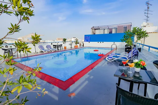 A rooftop swimming pool with red tile edging, striped sun loungers, small tables, and cocktail glasses on a table in the foreground.