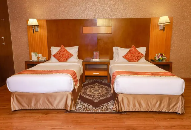 Two single beds with orange patterned cushions and orange-striped runners stand either side of a wooden nightstand in a hotel room with a patterned rug on a wood floor.