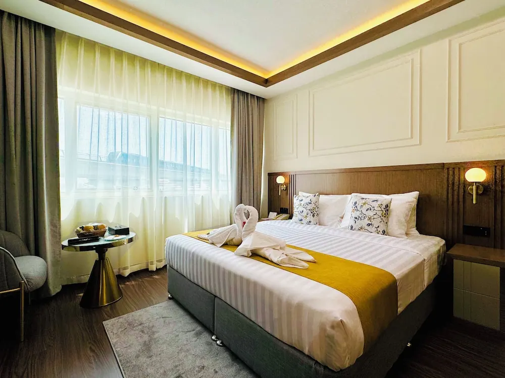 A hotel bedroom with a made king bed topped with a towel swan and a yellow runner, a round side table with a fruit basket, an armchair, and sheer curtains over a window.