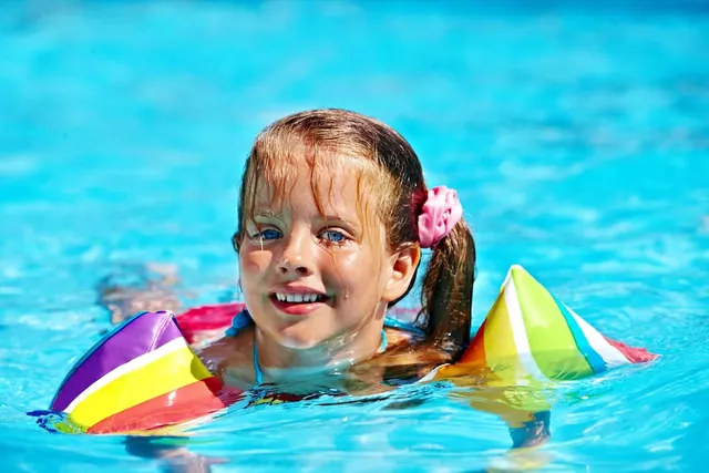 A smiling child with wet hair wears rainbow armbands while swimming in a bright blue pool.