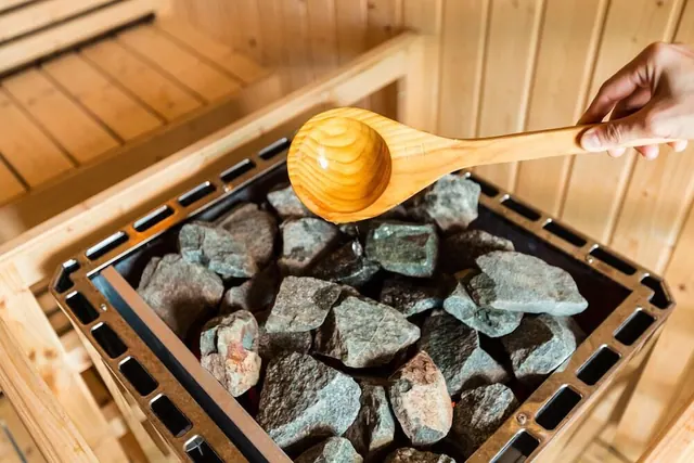 A hand holds a wooden ladle over stones in a sauna heater, with pine-panelled walls and benches behind it.