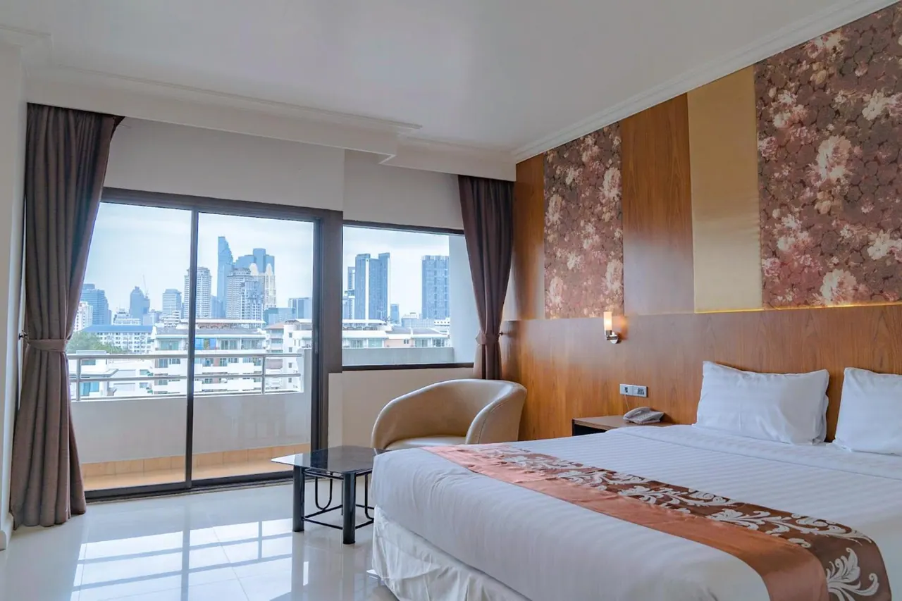 A bedroom with a large bed, an armchair and a glass side table, with sliding doors opening onto a balcony with a view of city high-rises.