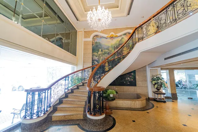 A curved staircase with ornate iron railings and a wooden handrail rises beside a painted mural and a crystal chandelier in the hotel lobby.